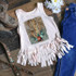 Leopard Cactus Sublimation Transfer, a white sleeveless top with fringe details and a leopard print cactus design, displayed on a rustic wooden surface with denim jeans, cowboy boots, and flowers.