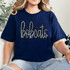 Bobcats Sequin Patch, a sparkly embroidered patch with the word 'bobcats' in cursive script on a navy blue T-shirt worn by a woman.