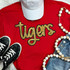Tigers Gold Sequin Patch, close-up view of a decorative golden sequin embroidered patch featuring the word 'tigers' on a red fabric shirt, with accessories including black sneakers, white cap, wooden bead necklace, and denim jeans.