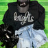 Knights Sequin Patch, a sparkling black patch with silver sequins shaped as a knight's helmet, displayed on a black sweatshirt with the word 'knights' in glittery script, laid out with denim shorts on a green textured surface.