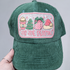 Tis The Season Embroidered Glitter Hat/Pocket Patch, green corduroy baseball cap with festive holiday-themed embroidered patch featuring a coffee cup, Christmas tree, gift box, and Santa hat, with glitter accents and pink embroidery.