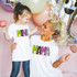 Mama Halloween Letters DTF Heat Transfer, a woman and a girl wearing matching white shirts with colorful 'MAMA' and 'MNI' text, celebrating with balloons and festive decorations.
