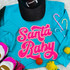 Santa Baby Sequin Patch, bright pink glittery patch with the words 'Santa Baby' in a playful script font on a turquoise sweatshirt.