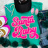 Santa Baby Sequin Patch, pink and white glitter patch with the text 'Santa Baby' on a teal T-shirt, paired with sneakers, jeans, and pink jewelry, decorated with green pom-pom garland.