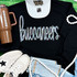 Buccaneers Sequin Patch, colorful glittery patch on a black t-shirt with casual accessories including white sneakers, a white cap, a brown tumbler, a pearl necklace, and denim shorts.