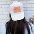 Jesus Loves You & I'm Trying Colorful Retro DTF Heat Transfer, displayed on a white trucker hat worn by a person with long brown hair against a white brick wall background.