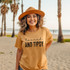 Minimalist Tanned And Tipsy BLACK DTF Heat Transfer, a woman wearing a tan T-shirt with this text and a wide-brimmed hat standing on a beach with palm trees in the background.