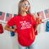 Give Me Liberty But Give Me Jesus First DTF Heat Transfer, woman wearing patriotic-themed sunglasses and a red T-shirt with the same text, holding a drink in a glass with strawberries, in front of American flags and decorations.