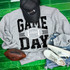 Game Day Various Colors/Puff Ink Screen Print, grey sweatshirt with black and white football design and text, paired with white sneakers, denim shorts, a black cap with glitter, and an American football on a green surface.