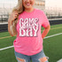 Pink Checker Game Day Coquette DTF Heat Transfer, pink t-shirt with a checkered game day design and the text 'Game Day' with a bow, on a woman at a sports field.
