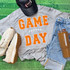 Game Day with Silicon Laces Patch - 10 Colors, sports-themed casual outfit with a gray T-shirt displaying 'Game Day' in orange lettering, a black cap, white sneakers, blue jeans, and a brown tumbler with a football-themed design.