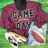Game Day with Silicone Laces Patch - 10 Colors, display of a maroon sweatshirt with 'Game Day' patch, pair of white and black sneakers, distressed denim shorts, and a football on a green turf background.