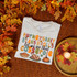 Thanksgiving Casserole DTF Heat Transfer, seen on a white t-shirt with colorful autumn-themed text and illustrations of a casserole dish, turkey, and pumpkin, placed on a textured orange blanket with fall leaves and a candle.