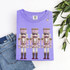 Hand-Painted Pastel Nutcrackers DTF Heat Transfer, three nutcracker design on purple t-shirt with a pair of blue jeans and a necklace, surrounded by green leaves.