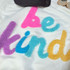 Be Kind Multicolor Fluffy Chenille Patch, colorful fuzzy embroidered patch with the words "Be Kind" in pink, blue, purple, and yellow on a white fabric background.