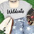 Wildcats Fluffy Chenille Patch Transfer | Varsity Game Day Patch, close-up of athletic apparel and accessories including a gray 'Wildcats' t-shirt, a beige 'Game Day' cap, jeans with stars, and baseball gloves and shoes on a grassy background.
