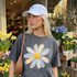 Faux Fabric Patchwork Daisy DTF Heat Transfer, a close-up of a woman wearing a dark gray t-shirt with a large, colorful daisy patchwork floral design on the front, in a flower shop with various flowers in the background.