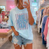 Big Jesus Fan Spangle Transfer, a woman wearing a light blue t-shirt with a glittery 'Big Jesus Fan' design and distressed denim shorts, taking a mirror selfie inside a clothing boutique.