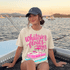 Whatever Floats Your Boat DTF Heat Transfer, vibrant pink boat graphic on beige t-shirt with text, worn by a woman on a boat during sunset