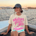 Whatever Floats Your Boat DTF Heat Transfer, woman sitting on a boat at sunset wearing a T-shirt with a pink boat graphic and the slogan 'Whatever floats your' in bright pink lettering.