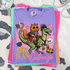 This Is An Outrage Retro Fantasy Cat DTF Heat Transfer, colorful digital graphic of a cartoon cat with sunglasses riding a dinosaur surrounded by mischievous elements like skulls, treasures, and potions.