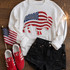 Patriotic Buffalo Sequin Patch, stylish casual outfit with American flag-themed buffalo patch on a white sweatshirt, paired with black distressed shorts, red Adidas sneakers with white stripes, an American flag, and a sparkler on a wooden background.