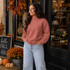 Blank Mockneck Sweatshirt, a woman wearing a cozy, rose-colored mockneck sweatshirt standing outside a boutique with fall decorations and multiple colors available.