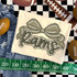 Camo Bow School Mascot DTF Heat Transfer, beige fabric with a camo bow design and the word 'Mom' in bold, decorative text, surrounded by footballs and a measuring tape for context.