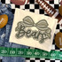 Camo Bow School Mascot DTF Heat Transfer, camouflage bow design with the word 'Bear' on a white fabric, surrounded by sports items including footballs and a measuring tape.