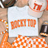 Rocky Top Football Laces Embroidered Chenille Patch, close-up of the patch on a gray sweatshirt featuring orange embroidered text 'ROCKY TOP' with laces design, surrounded by Tennessee Volunteers themed accessories including a hat, football, and themed decorations.