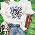 Game Day Sequin Patch With Football, casual sports-themed outfit with a white sweatshirt featuring a football and the words 'Game Day' in sequins, paired with blue jeans, white sneakers, a brown tumbler, and a pink wristband, laid out on a green football field background.