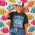 Game Day Polka Dot Helmet DTF Transfers | Football Color Combo Collection, a woman wearing a helmet-shaped polka dot design t-shirt and a colorful cap, standing in front of a wall decorated with football-themed smiley face patterns.