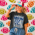 Game Day Polka Dot Helmet DTF Transfers | Football Color Combo Collection, a woman wearing a black T-shirt with sports-themed graphics and a colorful baseball cap with the text 'GAME DAY', standing against a background of football-shaped wall decorations with smiley face designs.