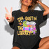Shh… Quiet in the Library DTF Transfer | Cute Librarian Bookworm Heat Transfer, a woman showing off a colorful and playful heat transfer design on a black t-shirt featuring a cartoon caterpillar, books, and stars with text 'Shh... Quiet in the Library!'