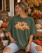 Hey There Pumpkin DTF Transfer, a retro fall script design on a green T-shirt worn by a woman in an autumn decorating setting with pumpkins and fall foliage.