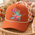 Dove Day DTF transfer featuring a whimsical patchwork-style dove in flight made of mixed bright patterns with bubble-letter DOVE and DAY text Shown on a Burnt orange front panel with cream rope and orange/ivory checkered mesh back trucker hat.