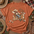 Cowboy swinging a lasso while riding Rudolick the reindeer DTF transfer, featuring western gear, studded saddle, and jingle-bell collar in illustrated style Shown on a Terracotta t-shirt.