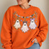 Pastel ghost garland DTF transfer with three bow-topped ghosts in orange, pink, and purple strung beneath a beaded candy-colored banner with stars and glitter accents Shown on a Orange crewneck sweatshirt.