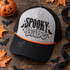 Spooky Babe Halloween DTF transfer with retro block lettering, graffiti-style script, bat silhouettes, sparkles, and checkerboard border on a mustard yellow shirt Shown on a heather grey front panel, black bill, black mesh back, orange rope trucker hat.
