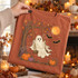 Cream-colored ghost swinging from a tree branch DTF transfer with glowing orange moon, bats, and autumn pumpkins Shown on a Terracotta t-shirt.