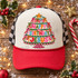 Happy Birthday Jesus DTF transfer featuring a folk-art Christmas tree-shaped cake with garlands, bows, a gold star topper, and rainbow lettering Shown on a cream front panel, red rope and bill, black-and-white checkered mesh back trucker hat.