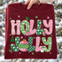Holly Jolly lettering DTF transfer in pink and green Christmas patterns with holly, gift box, stocking, ornament, tree, and snowman accents on a green shirt Shown on a Maroon crewneck sweatshirt.