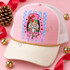 Holly Jolly nutcracker face DTF transfer with pink and gold design, red bow, green crown, and fluffy white beard in a pink scalloped medallion over a blue polka-dot striped background Shown on a blush pink front panel, ivory bill, white mesh trucker hat.