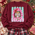 Holly Jolly nutcracker DTF transfer with pink and green nutcracker face, red bow, gold crown, in a pink scalloped frame over a mint polka-dot striped background with bold bubble lettering Shown on a Maroon crewneck sweatshirt.