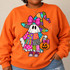 Patchwork ghost DTF transfer wearing a witch hat with pink bow, pink sneakers, and holding a jack-o-lantern bucket in a bold hand-drawn marker style Shown on a Orange crewneck sweatshirt.