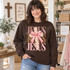 JESUS DTF transfer with pink leopard, giraffe print, and pink checkerboard lettering, layered with a wood-grain polka dot cross and pink gingham bow Shown on a Dark Chocolate crewneck sweatshirt.