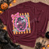 Cutesy Horror DTF Transfers | Pastel Spooky Icons Collection, burgundy T-shirt featuring a colorful cute horror character in a pink-framed design with stars, displayed among Halloween decorations