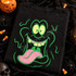 Goofy green monster face DTF transfer with bulging yellow eyes, yellow fangs, long pink tongue, and swirling green smoke squiggles on a black shirt Shown on a Black t-shirt.