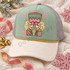 Embroidered-look nutcracker head DTF transfer with pink crown, green cap, star-shaped eyes, and cream fur beard surrounded by pink and sage snowflakes Shown on a sage front panel, cream bill, heather grey mesh, gold rope trucker hat.