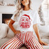 So Very Merry Santa DTF Transfer | Retro Christmas Design, red retro Santa face and “So Very Merry” lettering displayed on a white T-shirt worn with red-and-white striped pants in a cozy Christmas living room.